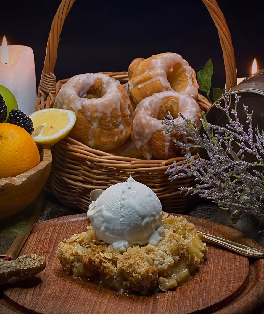 Apple crumble topped with a scoop of vanilla ice cream on a wooden plate, with a basket of glazed bundt cakes and assorted fruits in the background.