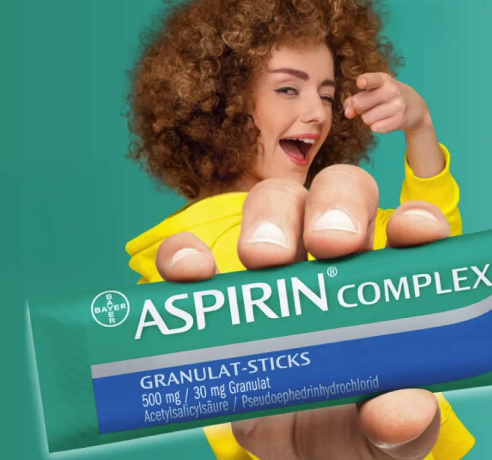 Frau mit lockigem Haar in gelber Jacke hält Aspirin Complex Granulat-Sticks vor einer grünen Wand.