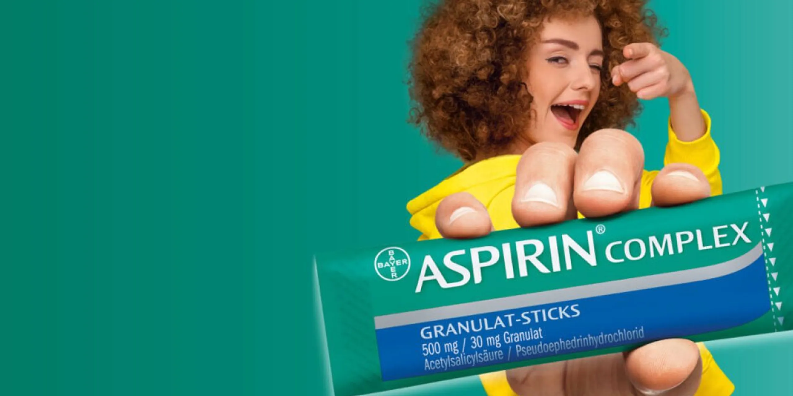 Frau mit lockigem Haar in gelbem Pullover zeigt und hält eine Packung Aspirin Complex Granulat-Sticks.