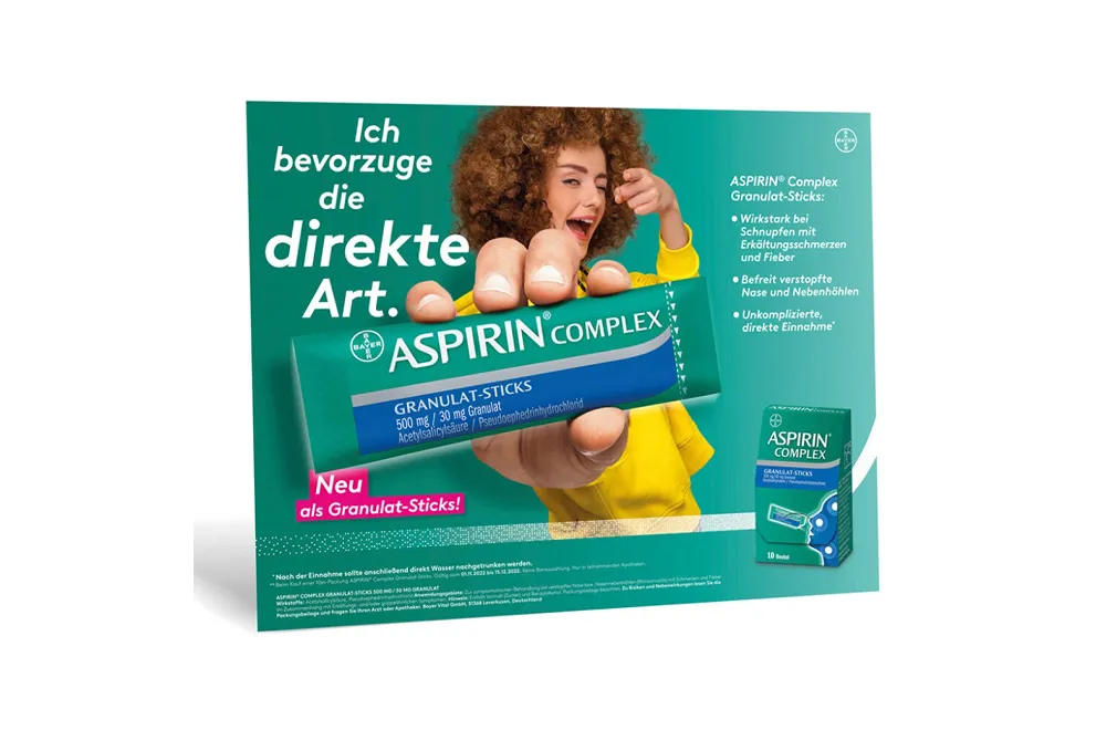 Frau hält eine Packung Aspirin Complex Granulat-Sticks vor, mit Text in Deutsch zur direkten Einnahme bei Erkältungsschmerzen und verstopfter Nase.
