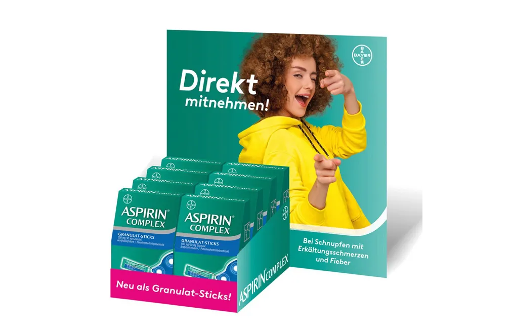 Display mit Aspirin Complex Granulat-Sticks und einer Frau im gelben Pullover, die direkt zur Kamera zeigt.