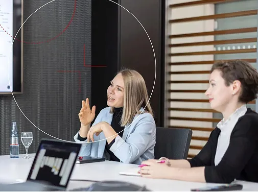 Zwei Frauen diskutieren bei einem Meeting in einem modernen Besprechungsraum, eine spricht und gestikuliert, die andere hört zu.