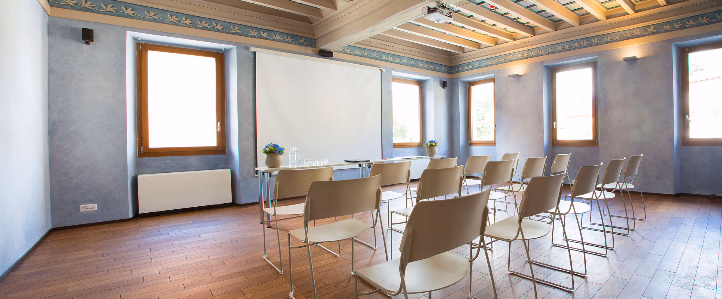 Heller Konferenzraum mit blauen Wänden, Holzböden, 15 weißen Stühlen und einem Tisch vor einer weißen Leinwand.