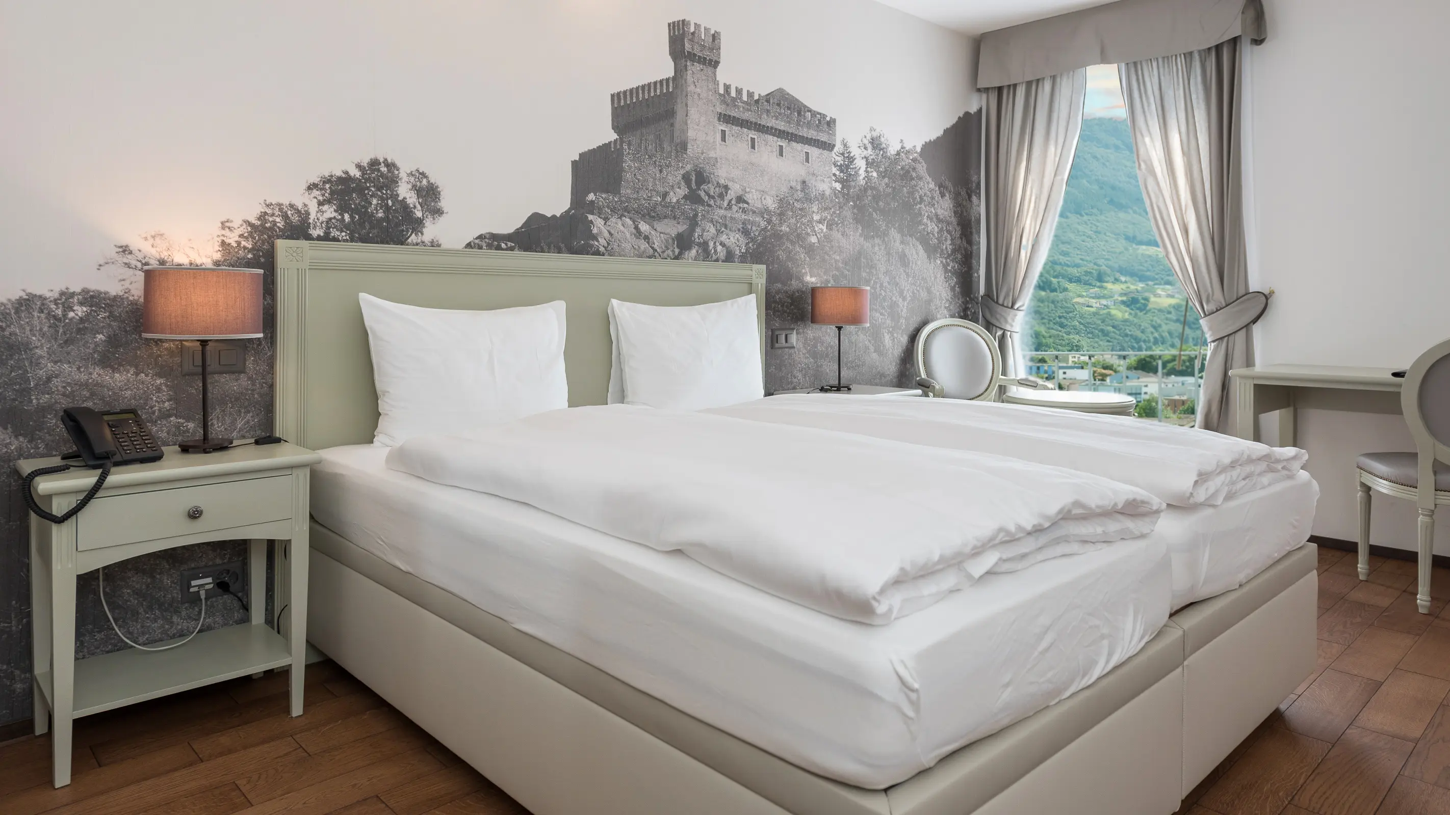Ampia e luminosa camera matrimoniale dell'Hotel La Tureta, caratterizzata da un suggestivo murale dei castelli di Bellinzona, pavimento in parquet e grande finestra con vista sulle montagne.