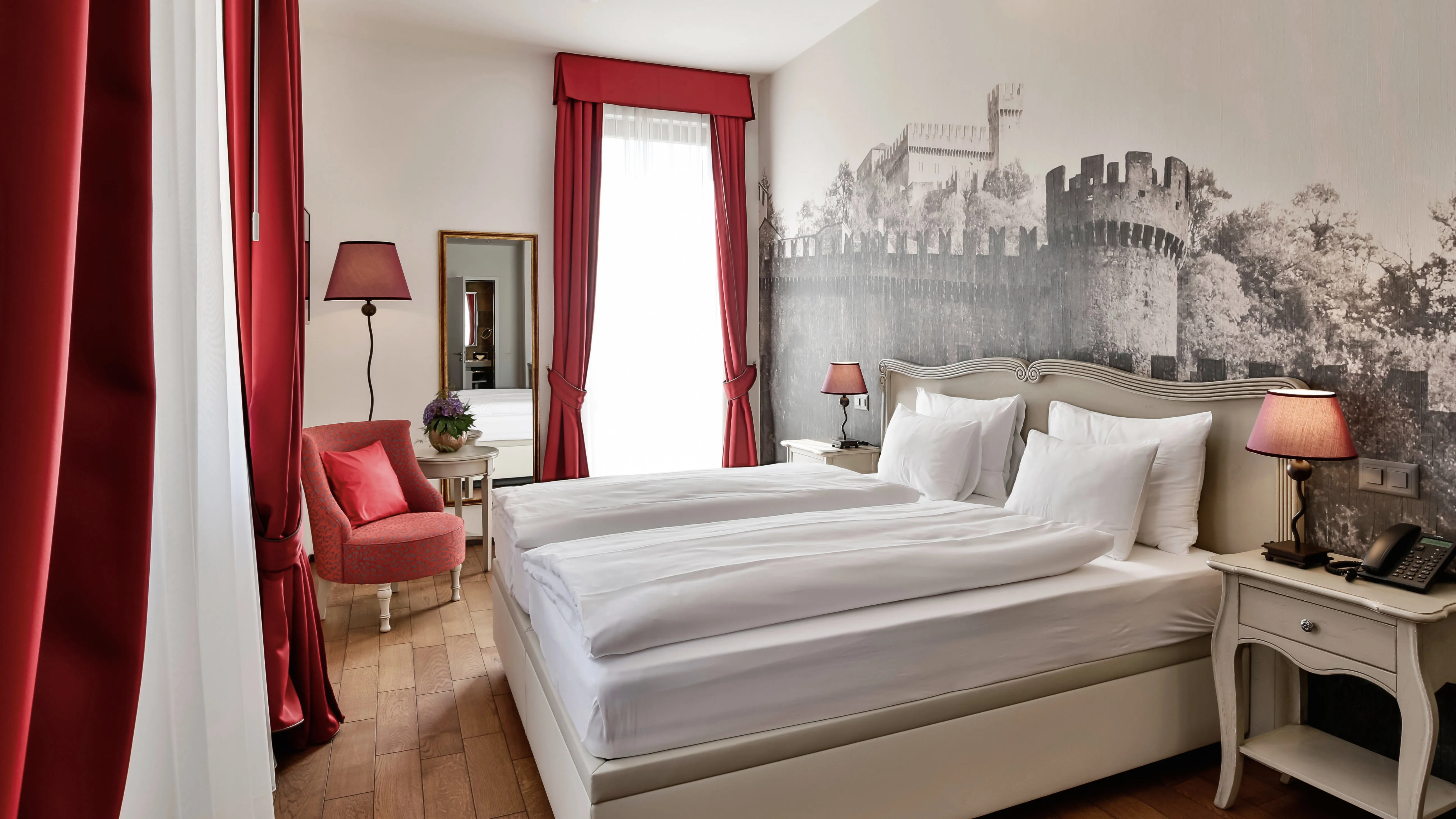 Accogliente Comfort Studio dell'Hotel La Tureta: letto matrimoniale incorniciato da eleganti tende rosse che filtrano la luce naturale, affiancato da arredi in stile classico e lampade abbinate.