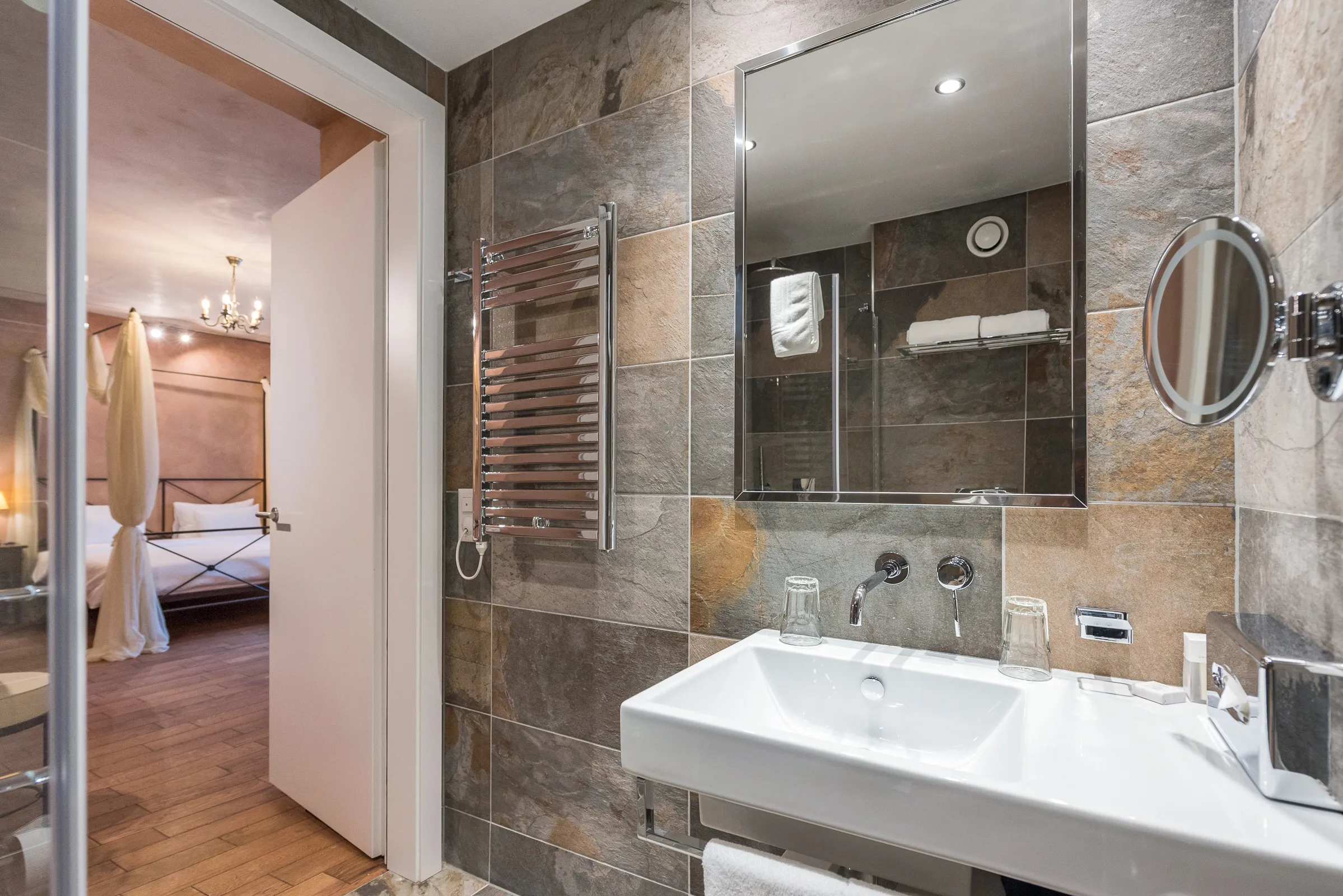 Moderno bagno privato della Camera Unique, rifinito con eleganti piastrelle effetto pietra; l'ampio specchio riflette la zona notte con il letto a baldacchino.