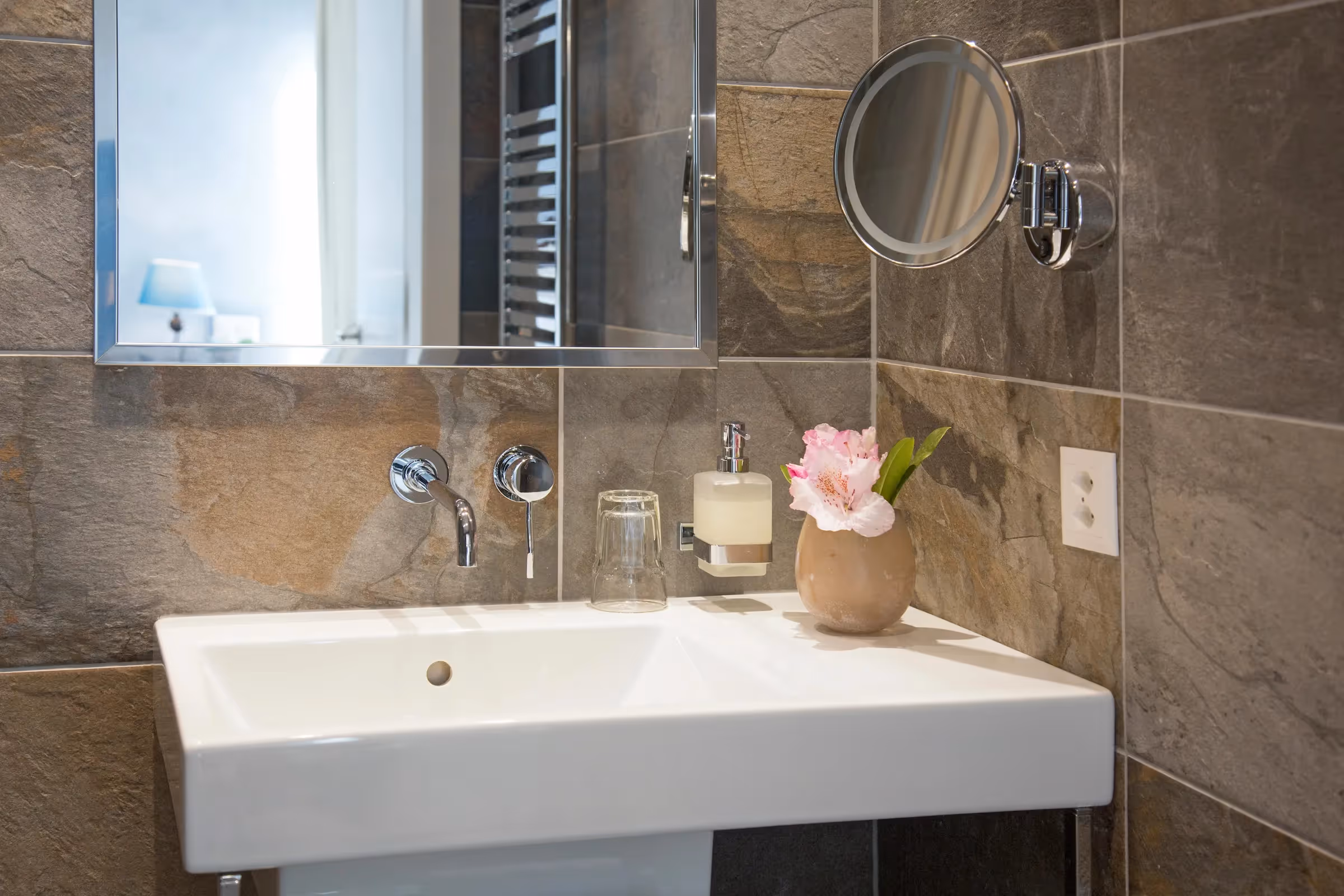 Dettaglio lavabo Business: Lavabo moderno bianco con rubinetteria a muro cromata, specchio ingranditore e rivestimento in pietra naturale multicolore.