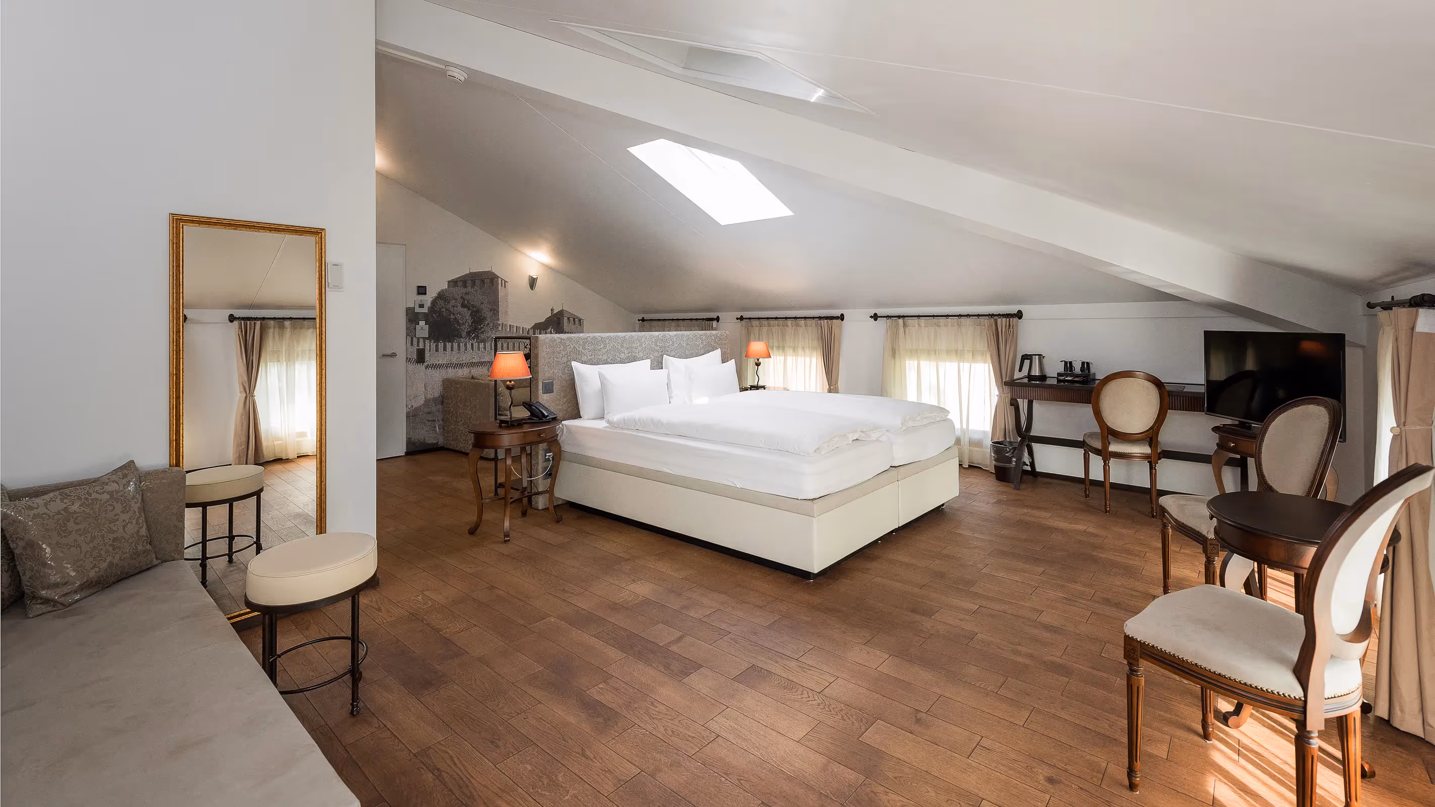 Vue panoramique de la Suite Design Loft : grande chambre mansardée avec lit double, canapé beige et coin bureau éclairé par une lucarne.