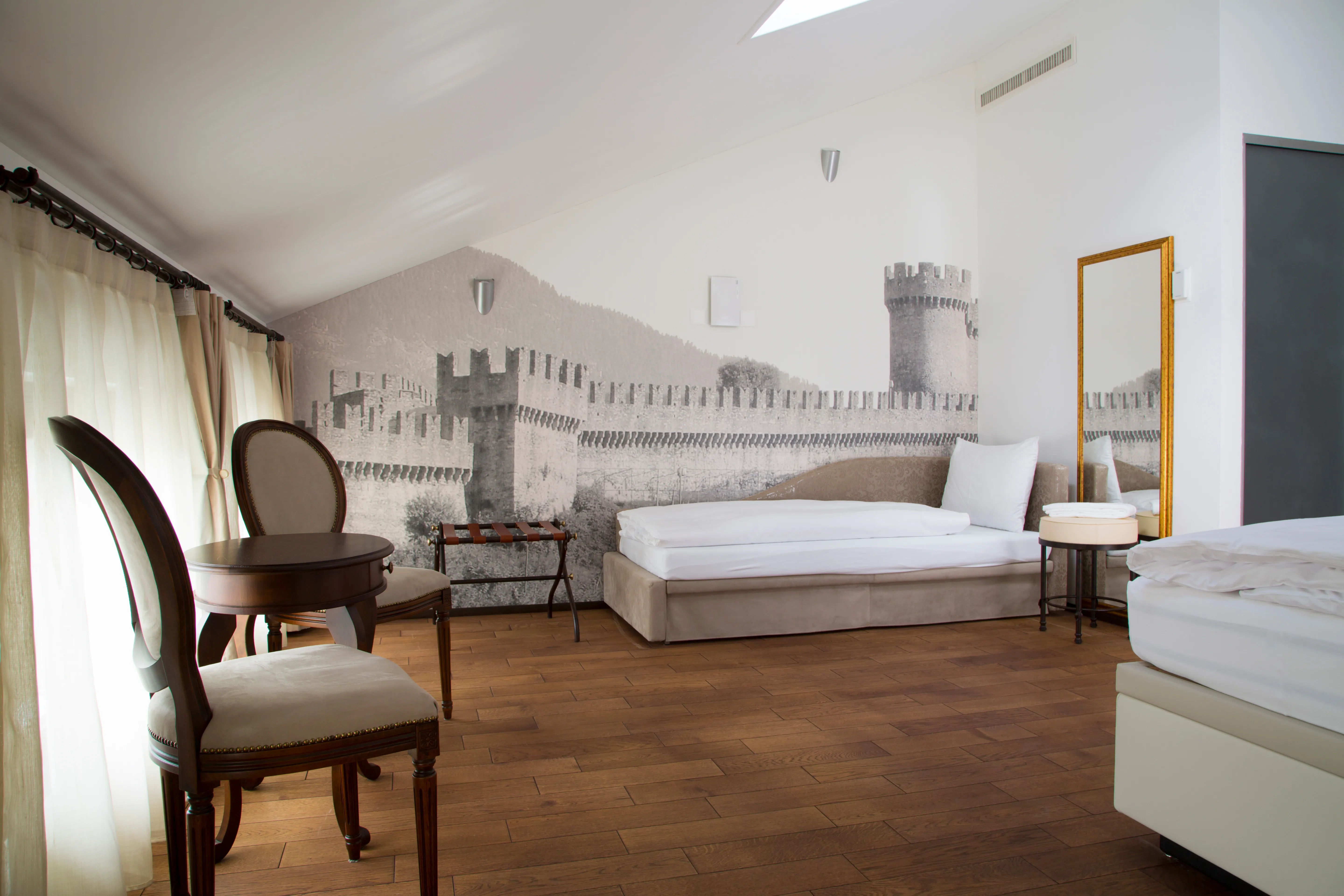 Zona giorno e divano letto: Parete decorata con un murale in bianco e nero raffigurante i castelli di Bellinzona. Include un divano letto beige e specchiera con cornice dorata.