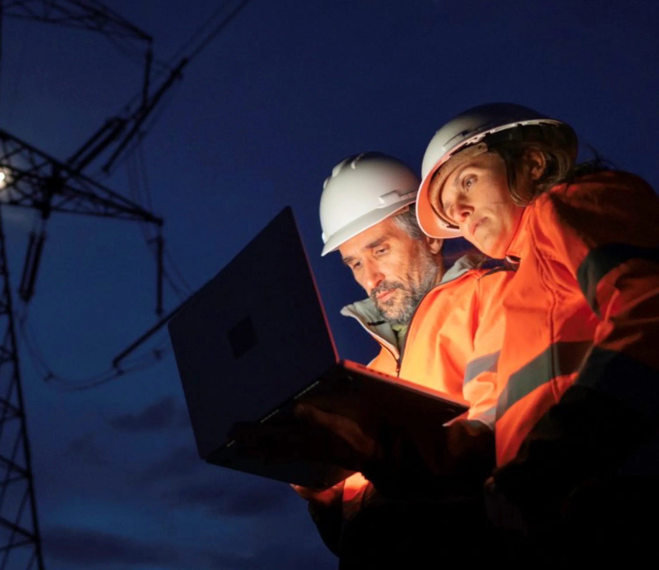 Two engineers in hard hats and orange safety jackets working on a laptop at dusk near electrical power lines.