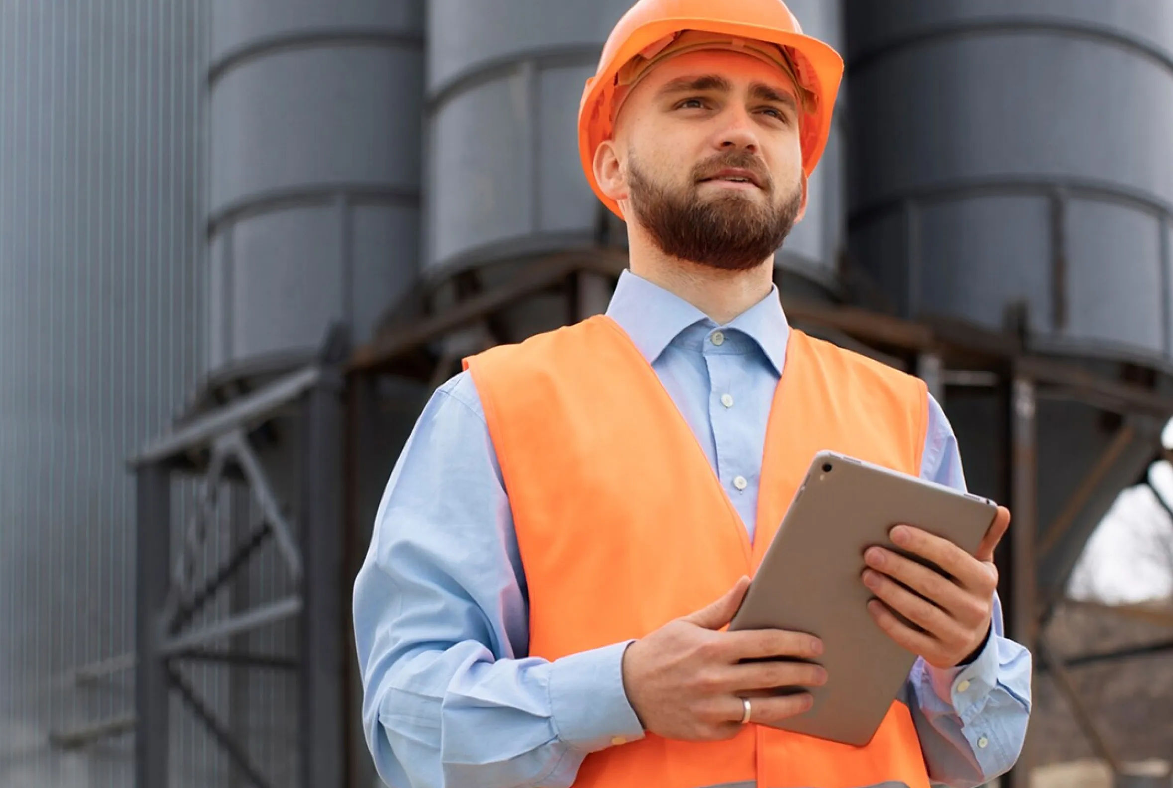 Bearded construction worker wearing an orange safety vest and helmet holding a tablet, standing near industrial silos.