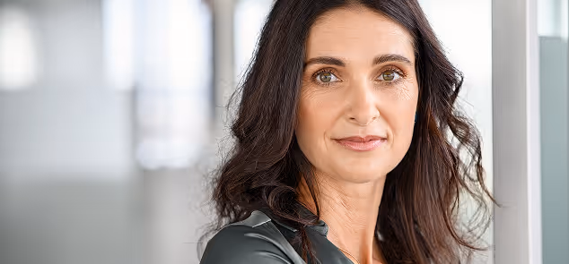 Confident middle-aged woman with dark hair and brown eyes standing indoors against a window.