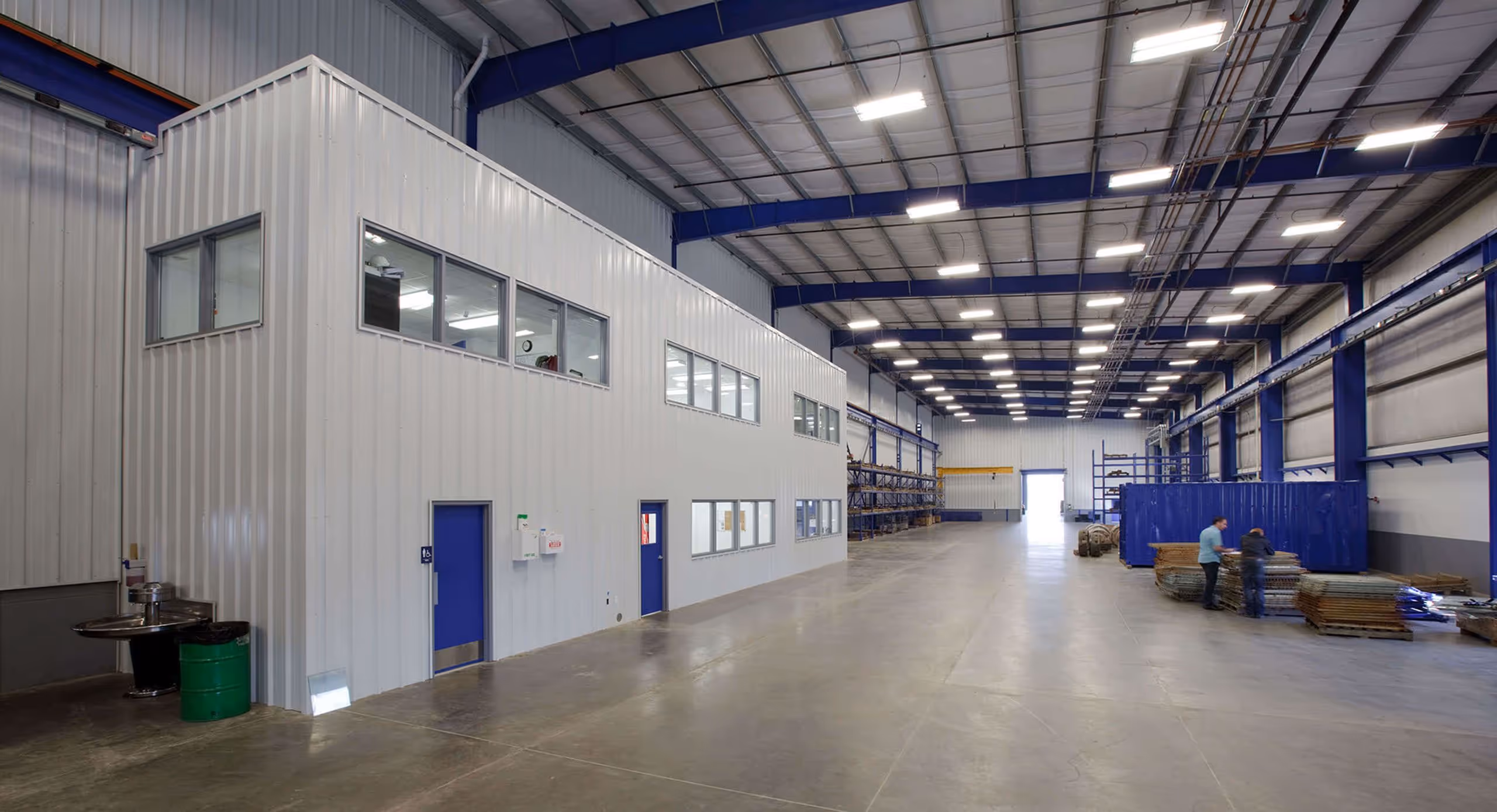 Spacious industrial warehouse interior with white office structure, blue doors, metal racks, and two workers near stacked materials.