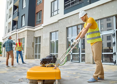 Construction worker in a safety vest and helmet operating a yellow floor cleaning machine outside a building.