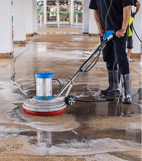 Person using a floor cleaning machine to scrub a wet floor in a spacious indoor area.