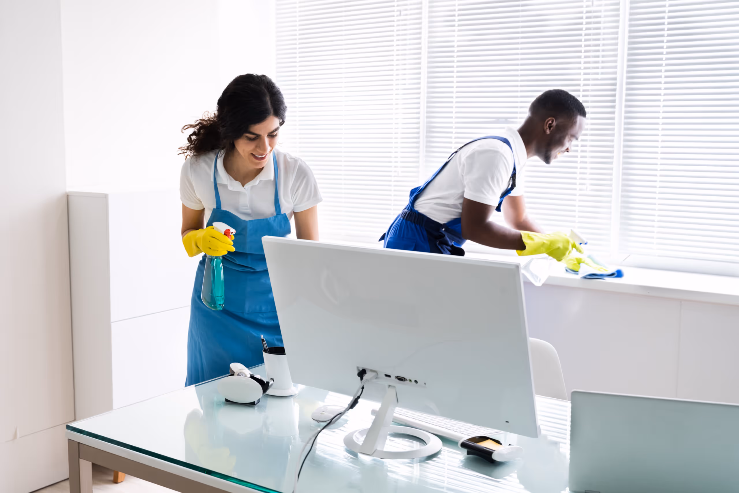 Two professional cleaners wearing blue aprons and yellow gloves cleaning a bright office with a spray bottle and cloth near a computer.
