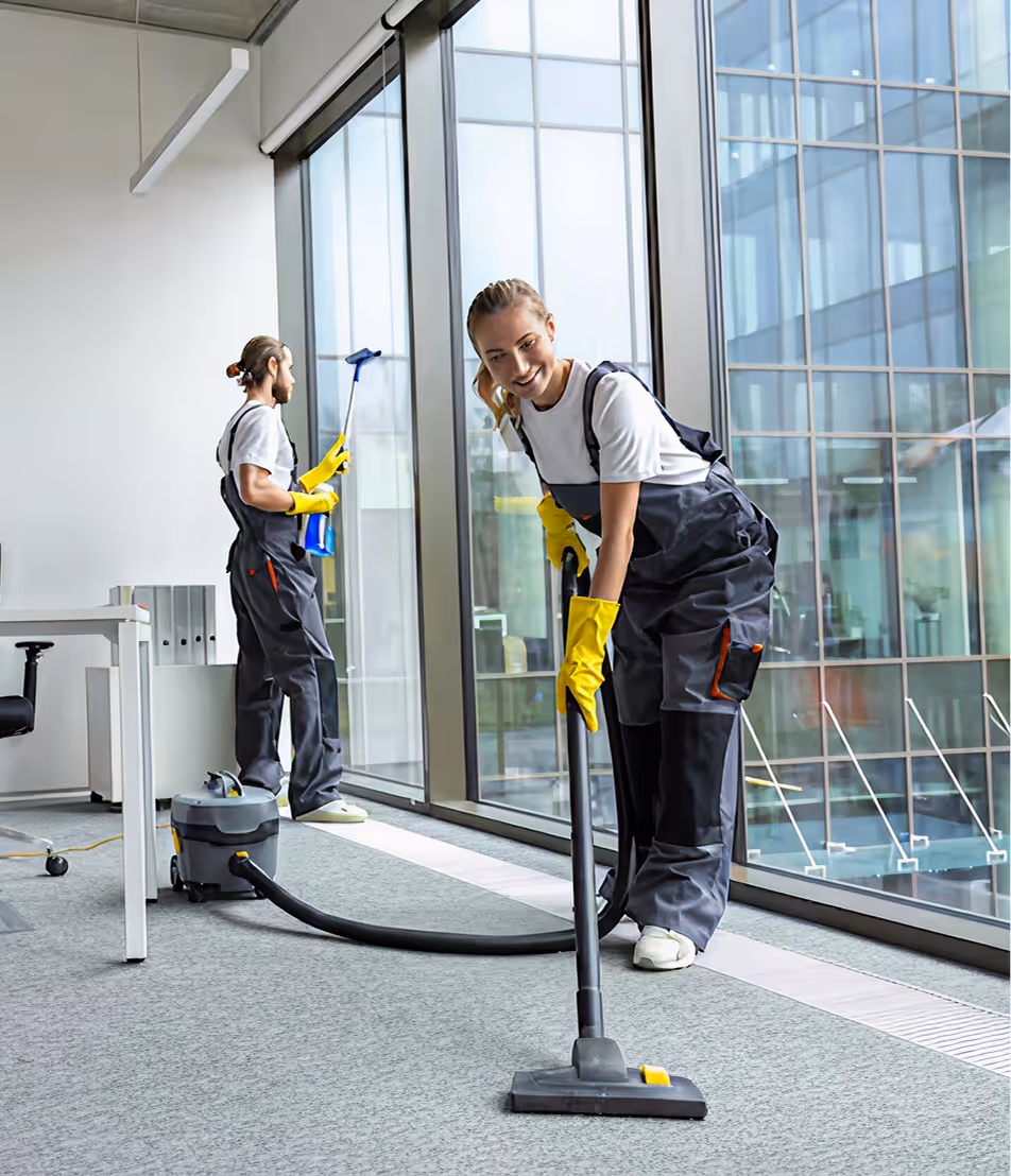 Two professional cleaners in overalls cleaning an office with large windows, one vacuuming the carpet and the other cleaning the glass.