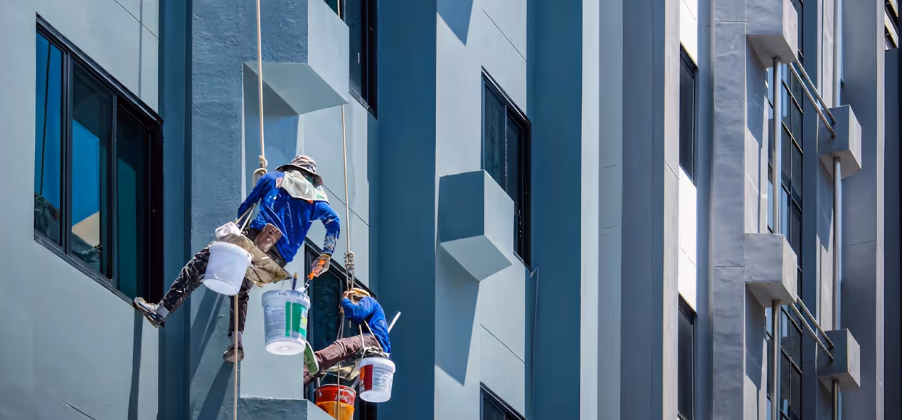 Two workers suspended by ropes cleaning or painting the exterior windows of a modern high-rise building.