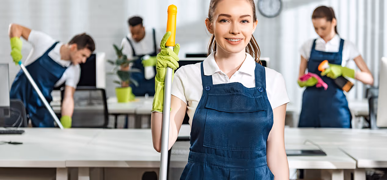 Smiling woman in cleaning uniform holding a mop in an office while colleagues clean desks in the background.