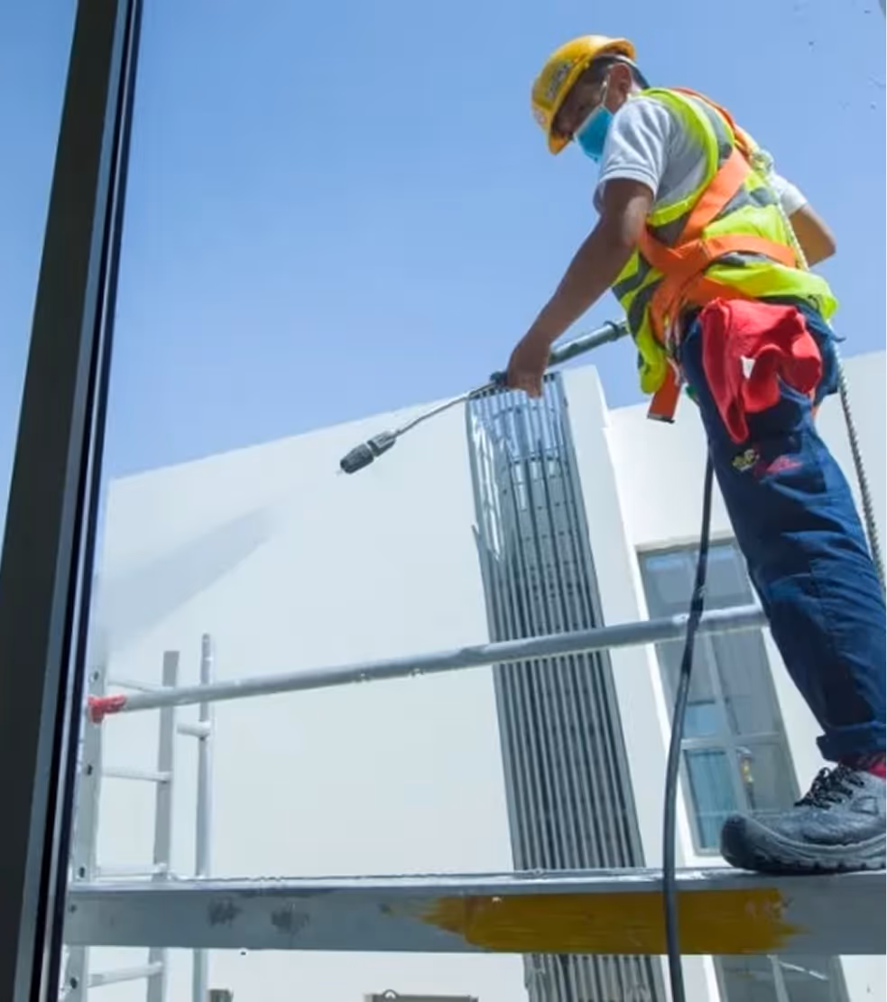 Construction worker wearing a yellow helmet and safety vest using a power washer on a building exterior.