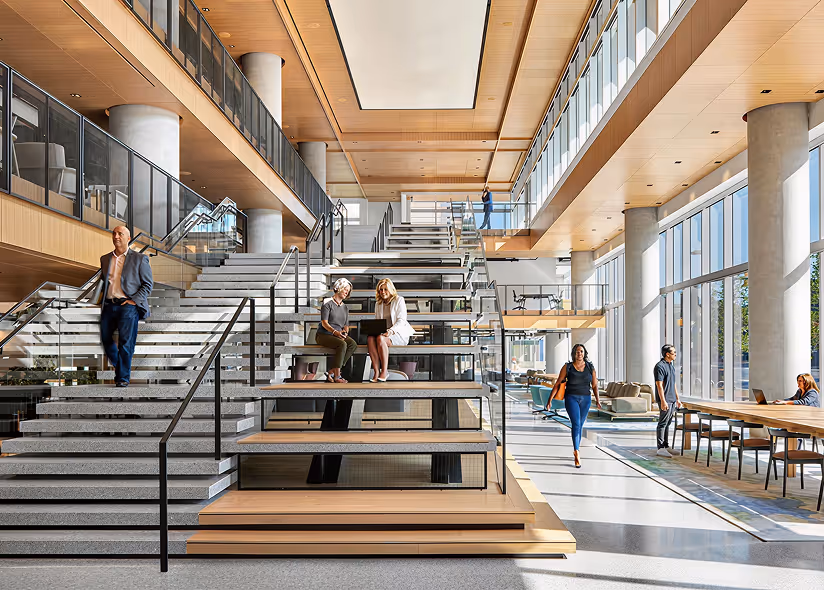Modern office interior with large windows, wooden ceilings, wide staircase, and people working and walking inside.