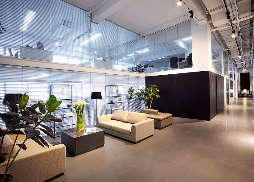 Modern office lobby with beige sofas, glass walls, indoor plants, and black shelving units.