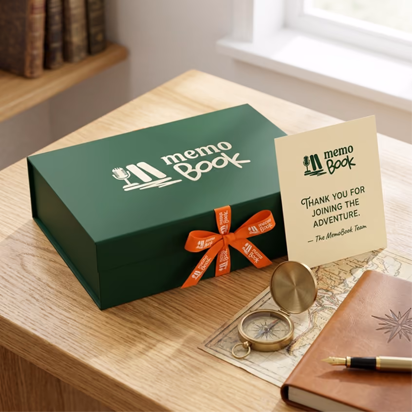 Green MemoBook gift box with orange ribbon, card, compass, map, and leather notebook with pen on a wooden desk.