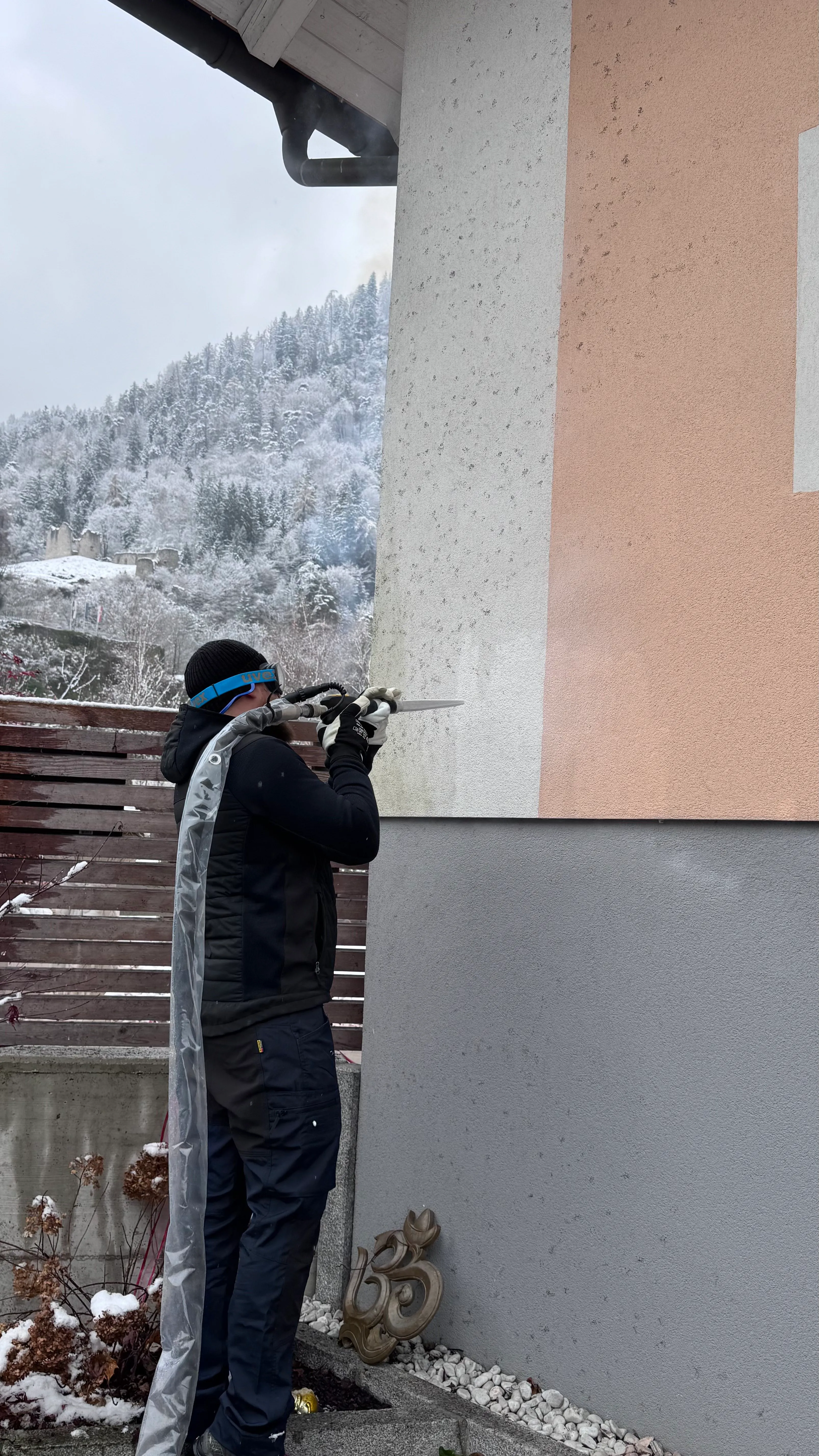Person hält Gerät mit Schlauch, sprüht Wand in einer verschneiten Berglandschaft.