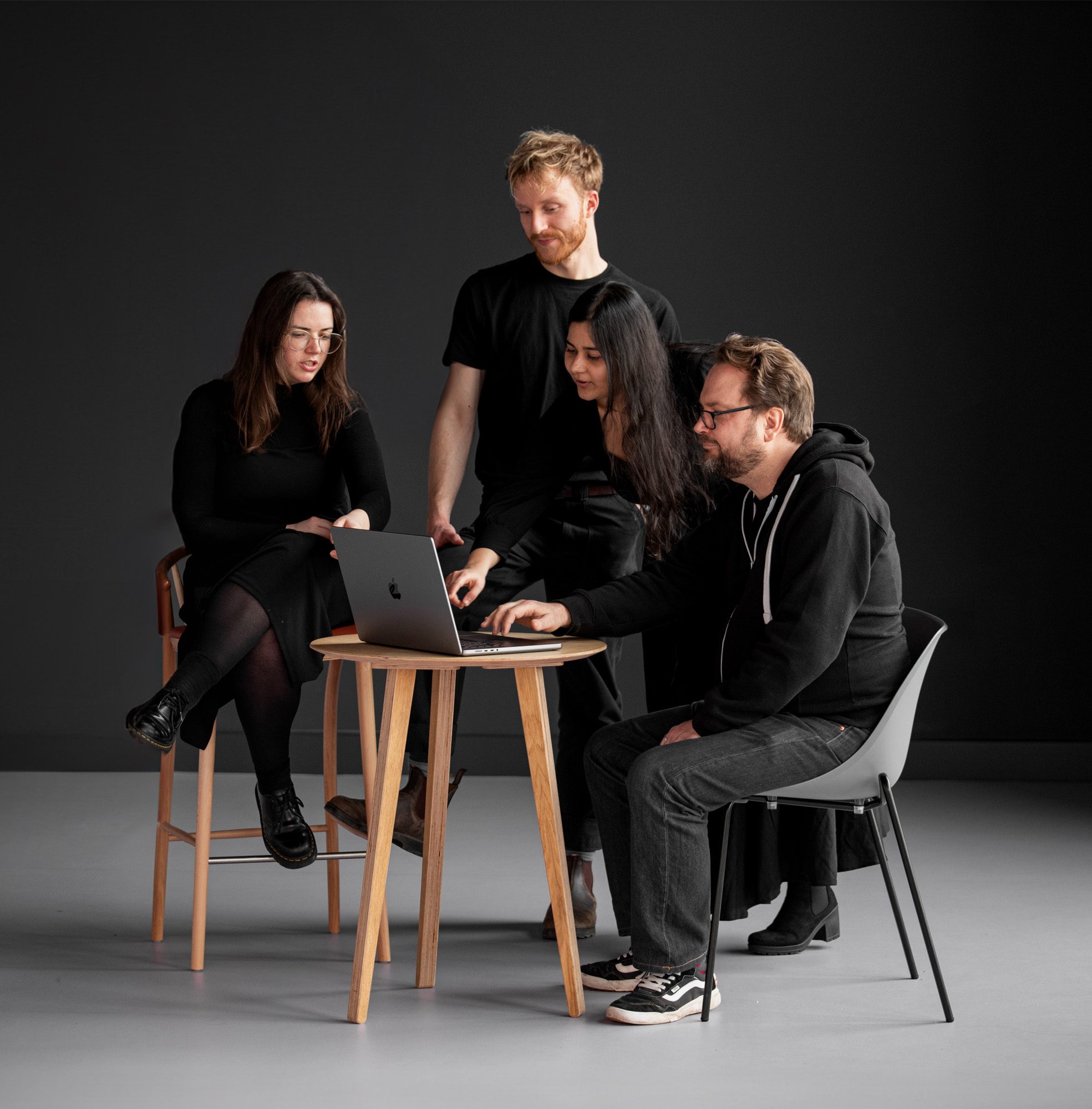 Four people gathered around a wooden table, looking at a laptop screen, in a minimal indoor setting with a dark background.