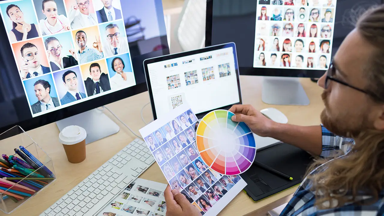 Man with glasses and long hair holding a color wheel and photo sheets while working at a desk with three monitors displaying various headshots.