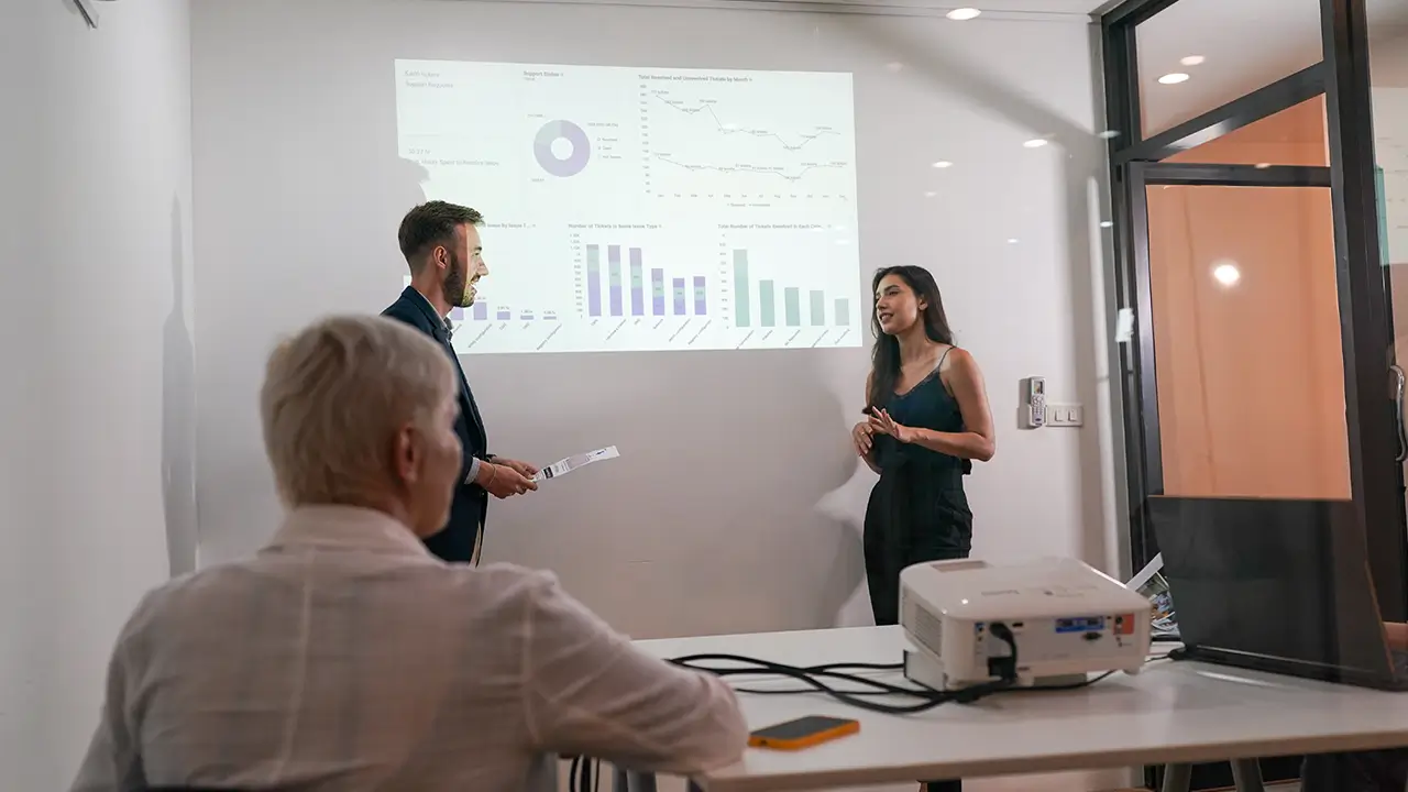 Two professionals presenting data charts projected on a white wall while a third person watches seated at a table with a projector.
