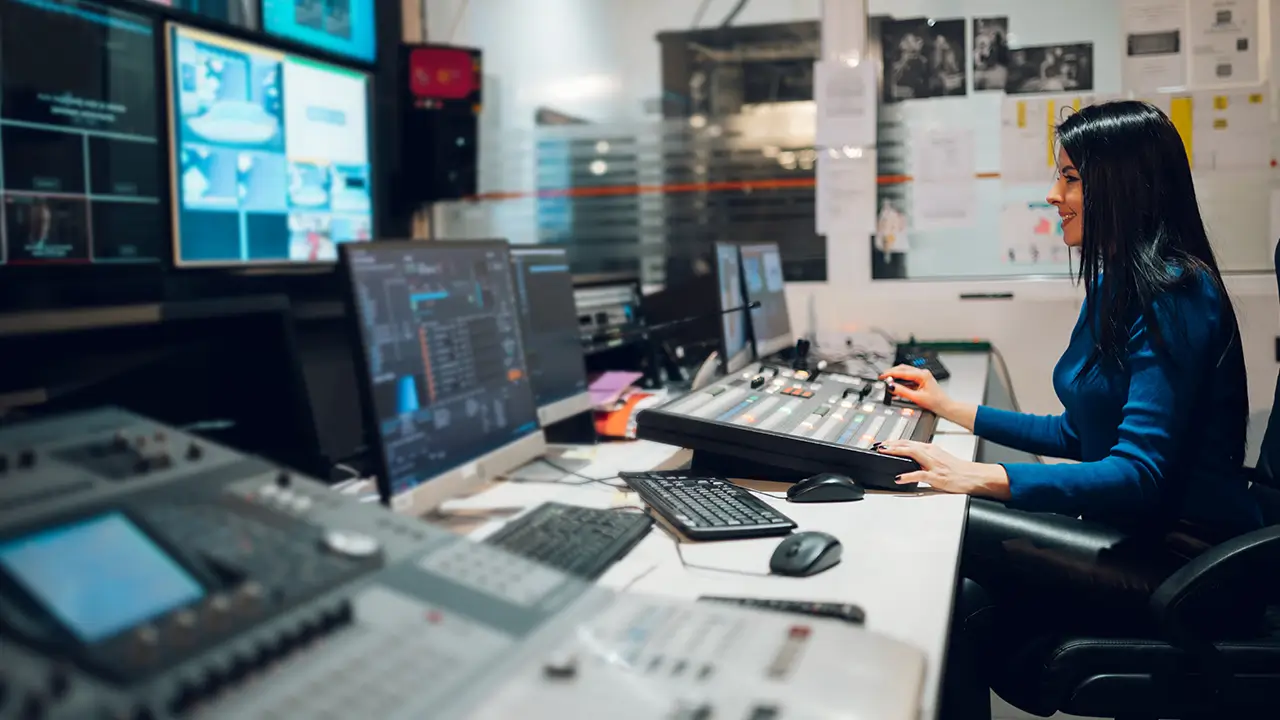Woman operating audio mixing console and multiple monitors in a control room.