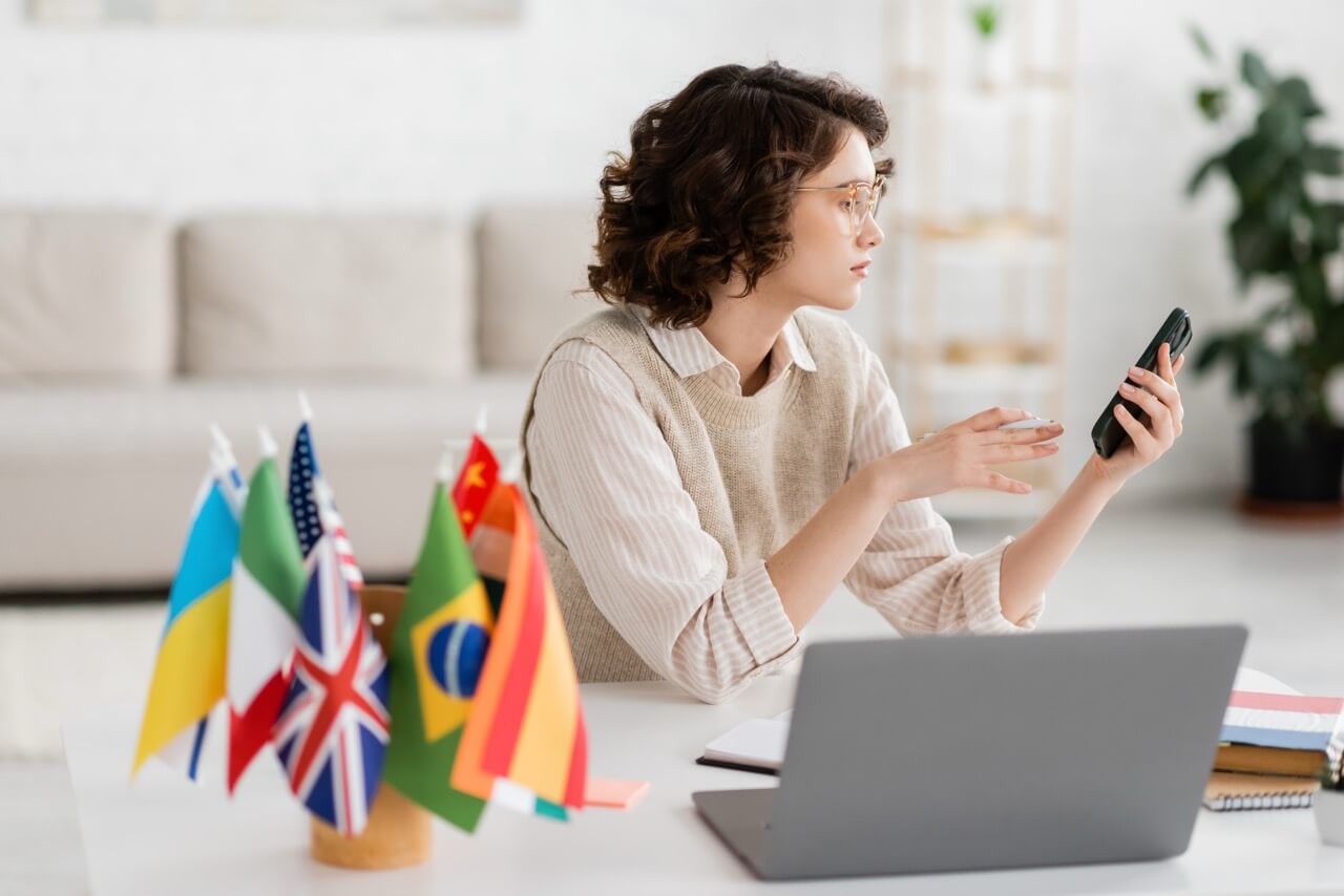 Young woman with glasses sitting at a desk with a laptop and several international flags, holding and looking at a smartphone.