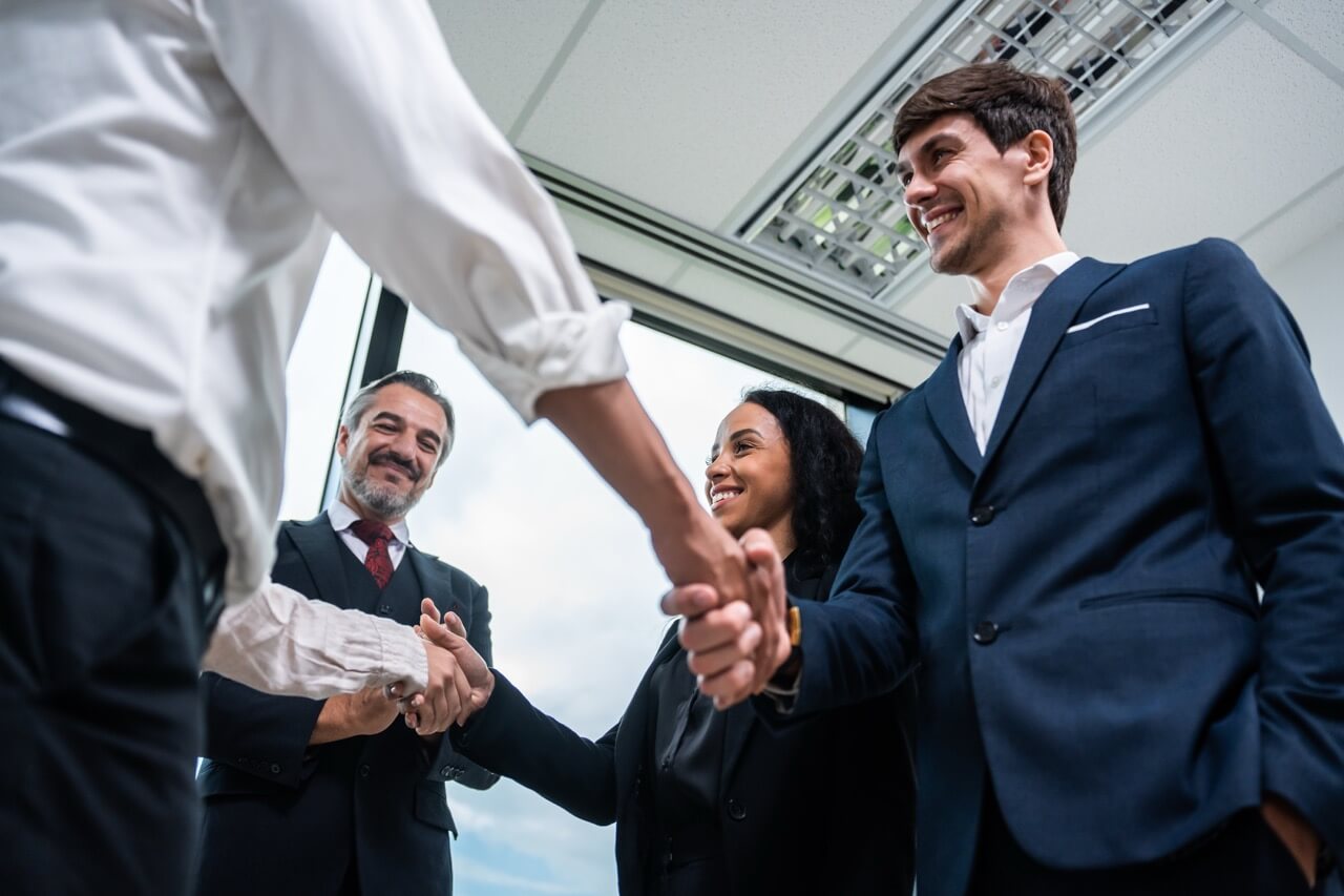 Four businesspeople in formal attire smiling and shaking hands in an office with large windows.