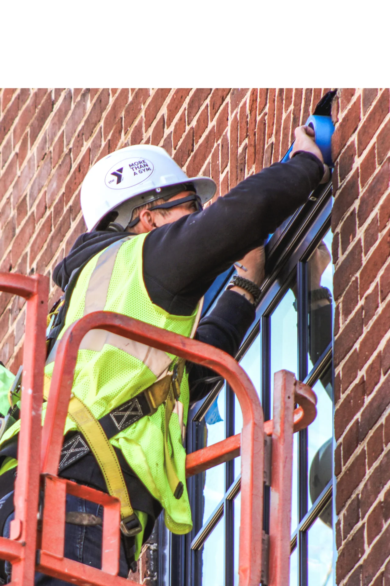 Construction worker wearing a safety vest and hard hat applying blue tape around a window on a red brick building from a lift platform.