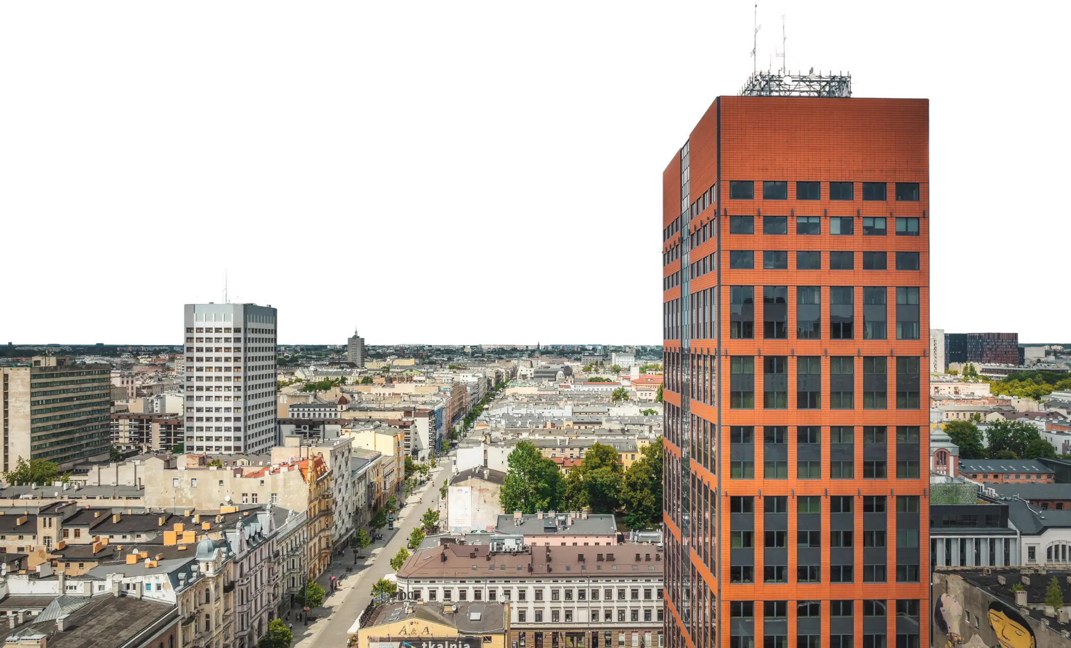 Widok na miasto z wieżowcem o czerwono-pomarańczowej elewacji na pierwszym planie i ulicą biegnącą w głąb panoramy.