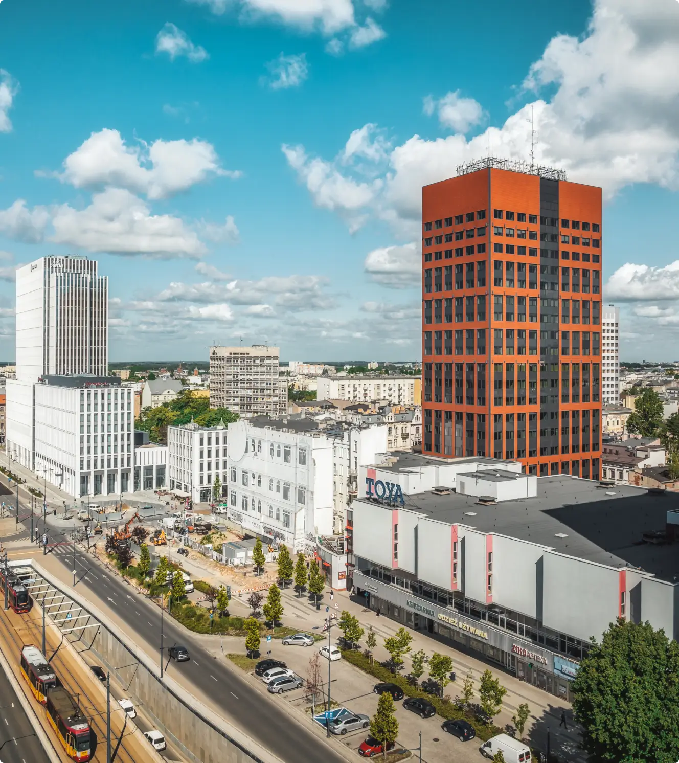 Panorama miasta z czerwonym wieżowcem, białymi budynkami, ulicą z tramwajami i parkingiem przy słonecznej pogodzie.
