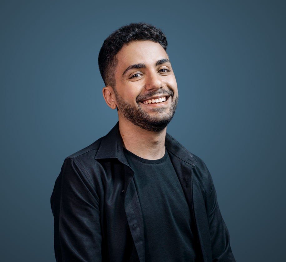 Smiling man with curly hair and beard wearing a black shirt and jacket against a blue background.