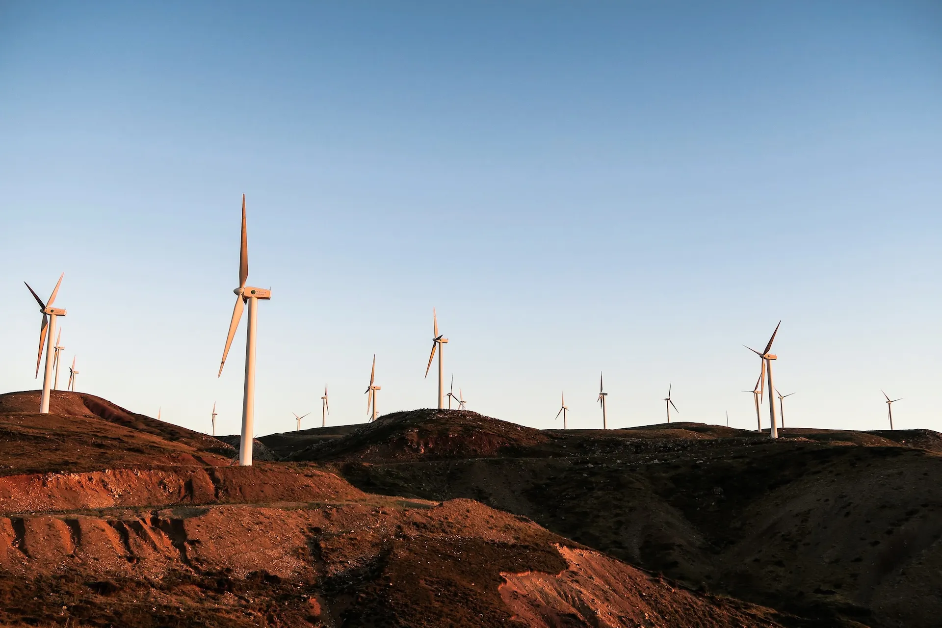 Wind turbines scattered across hills under a clear blue sky at sunset.