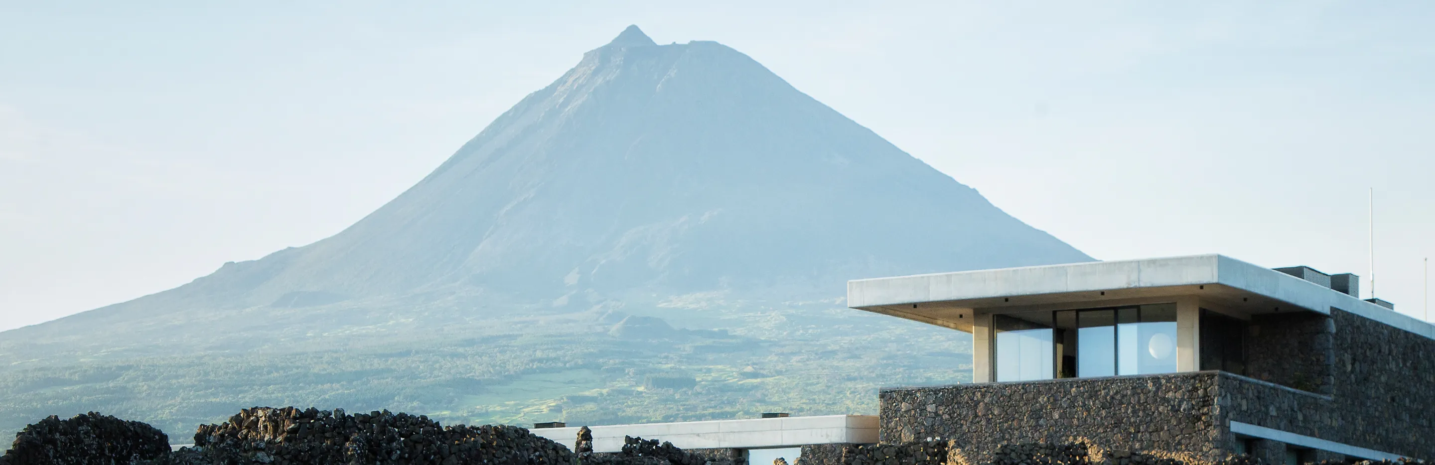 Modern stone building with large windows in front of a large green mountain under a clear sky.