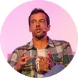Man with short hair wearing plaid shirt, speaking and gesturing on stage with microphone.