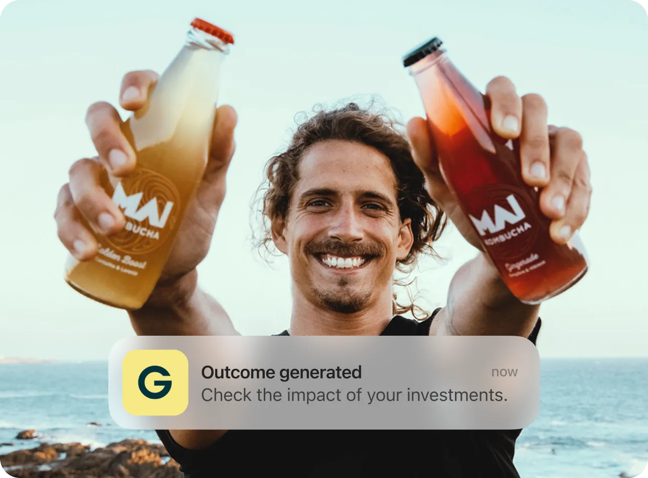 Smiling man holding two bottles of MAN Kombucha by the sea with rocks and ocean in background.
