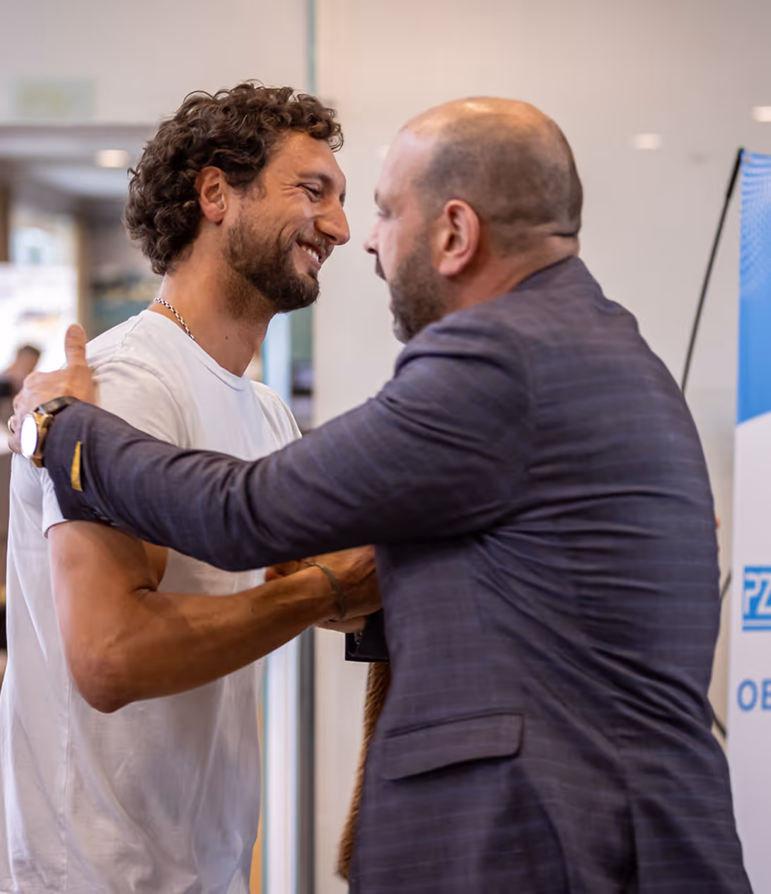 Two men greeting warmly with smiles and a handshake in an indoor setting.