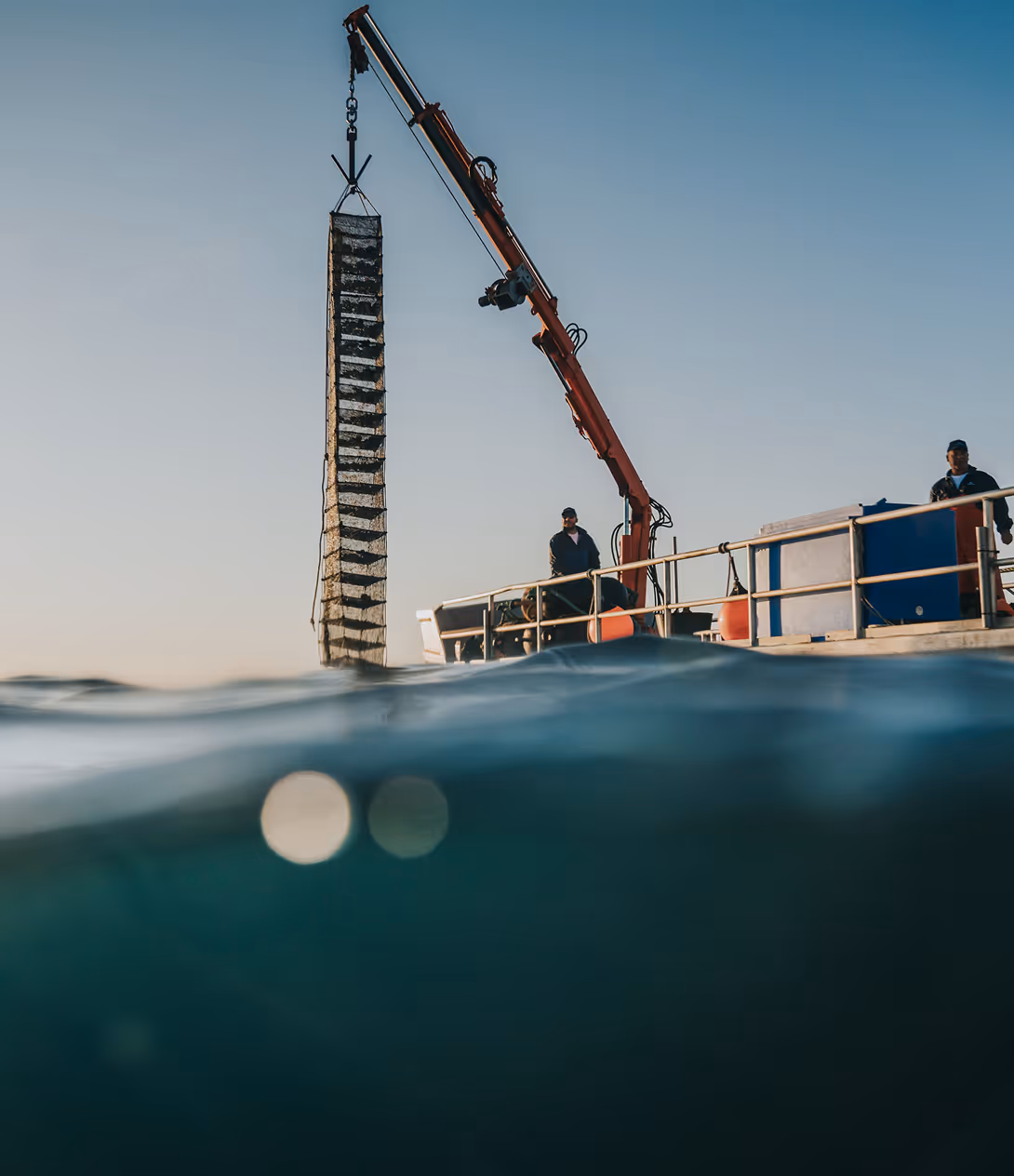 Boat crane lifting a tall, rectangular fishing cage from the water with two men on deck.