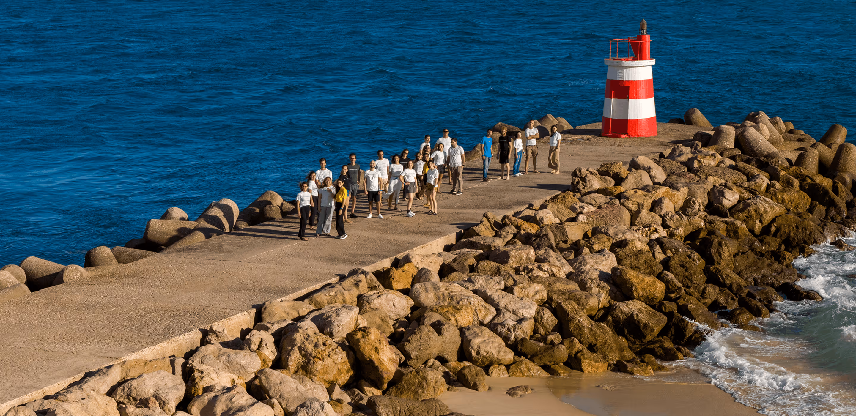 Group of people standing on a rocky pier near a red and white striped lighthouse by the ocean.
