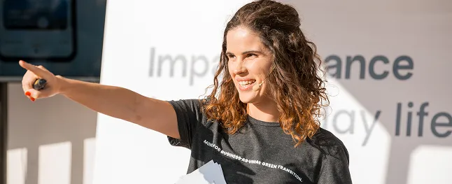 Smiling woman with microphone points while holding paper during a presentation.