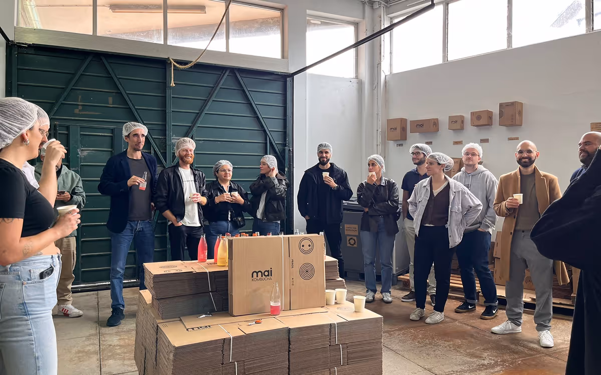Group of people wearing hairnets standing in a warehouse, drinking beverages near stacked cardboard boxes.
