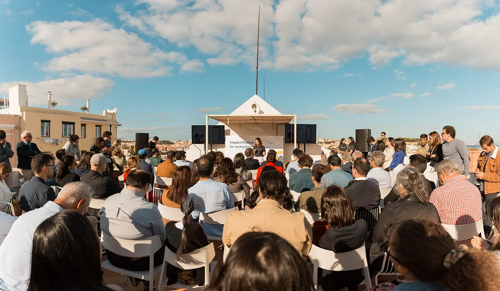 Audience seated outdoors listening to a speaker in front of a sign reading Impact finance for everyone.