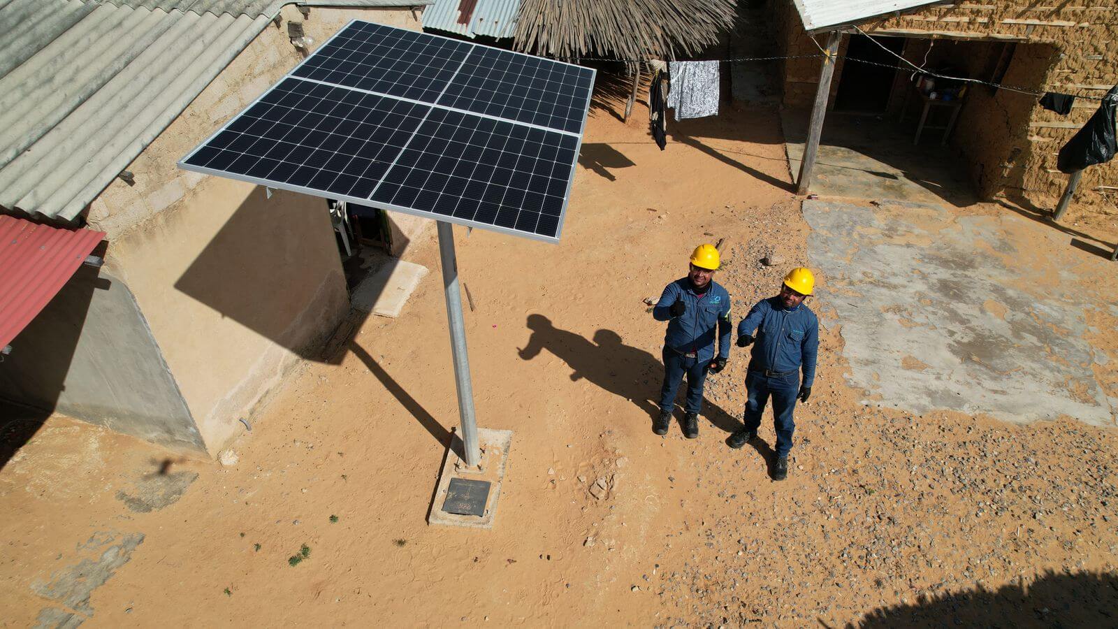 Two workers in yellow helmets giving thumbs up next to a ground-mounted solar panel in a rural area.