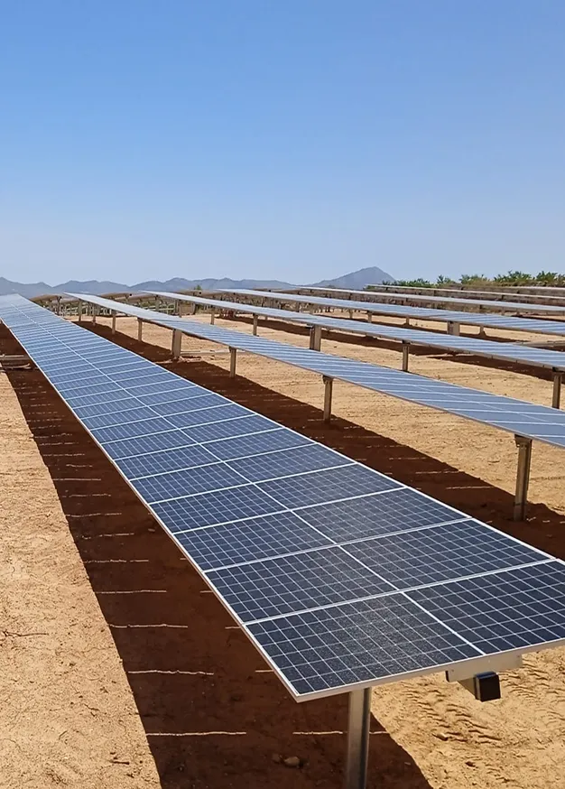 Rows of solar panels installed on metal supports in a sunny, arid landscape with mountains in the distance.